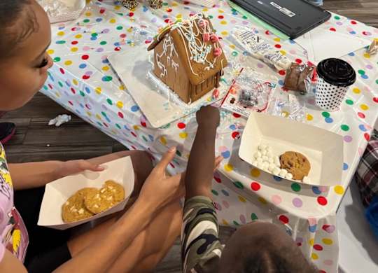 A person is sitting at a table with a gingerbread house and cookies.
