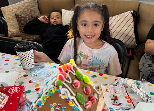 A young girl is sitting at a table with a gingerbread house in front of her.