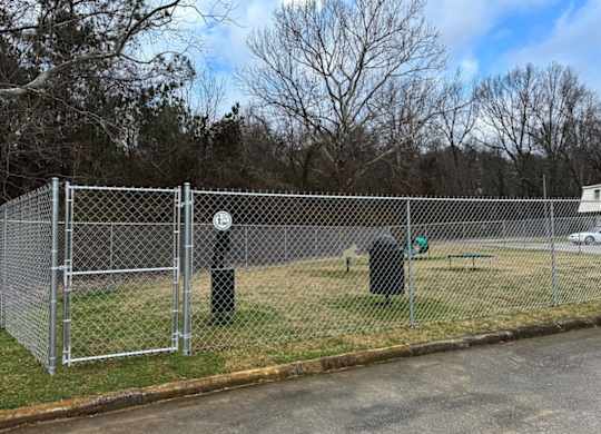 A silver chain link fence surrounds a green trash can and a green bench.
