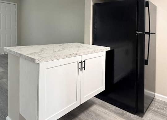 A black refrigerator stands next to a white cabinet with a marble top.