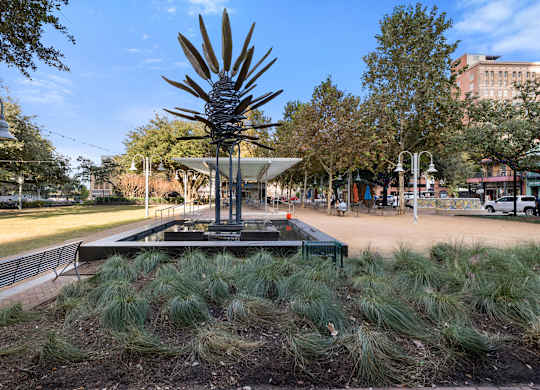 A large metal plant sculpture is the centerpiece of a park with a bench and a fence in the foreground.