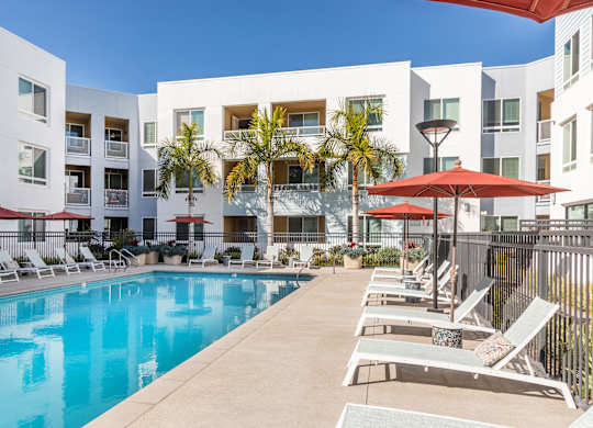 A swimming pool surrounded by lounge chairs and umbrellas in front of a white apartment building.