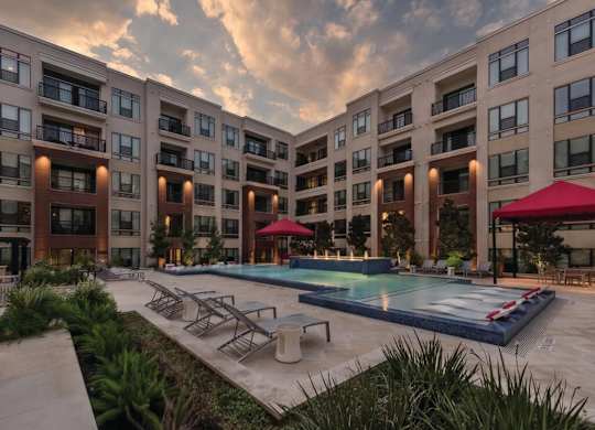 A large swimming pool is surrounded by lounge chairs and umbrellas in front of apartment buildings.