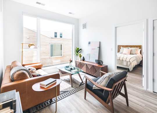 A living room with a brown sofa, a glass coffee table, and a bed in the background.