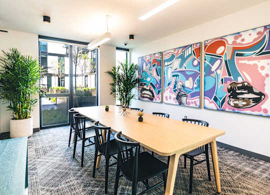 A conference room with a long table and chairs, a potted plant, and a colorful mural on the wall.