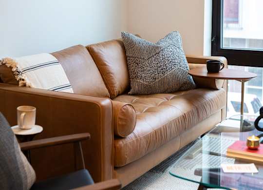 A brown leather couch with a grey pillow and a small table with a cup on it.