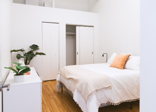 a bedroom with white walls and a white bed with an orange pillow at The Ferguson Apartments, Detroit, 48226