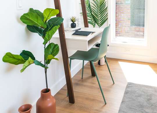 A room with a wooden floor, a green chair, a wooden ladder, a desk with a laptop, and a potted plant.