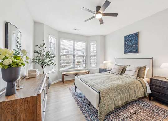 Luxury Apartments in Chapel Hill, NC, With In-Unit Washer and Dryer - Reserve at Meadowmont - Bedroom with Wood-Style Flooring, Bed, Large Wooden Dresser, Two Nightstands with Lamps, Ceiling Fan, and Large Windows with Blinds.