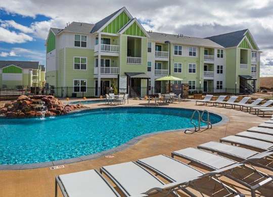 A large swimming pool surrounded by sun loungers in front of a multi-story apartment building.