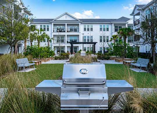 A silver grill is in the foreground of a grassy area in front of a white building.