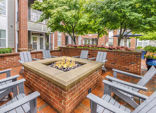 3-Bedroom Apartments in Charlotte, NC - Gramercy Square at Ayrsley - A Square Brick Fire Pit Surrounded by Gray Adirondack Chairs, Low Brick Wall, and Trees.