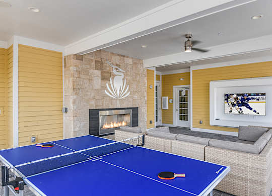 A blue ping pong table is in the middle of a living room with a fireplace and a painting on the wall.