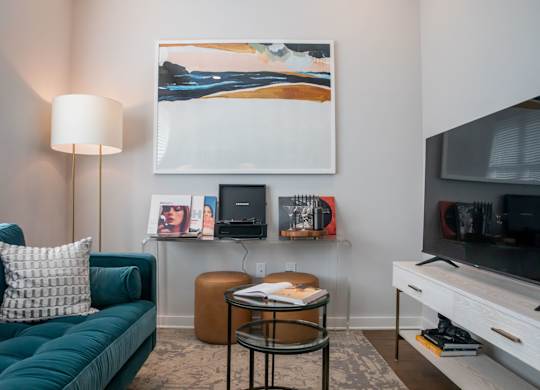 A living room with a green couch, a table with a lamp, a painting on the wall, and a television.