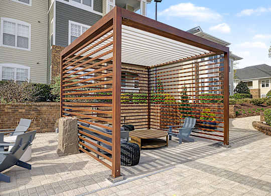 A wooden pergola with a metal roof is situated on a patio with a bench and chairs.