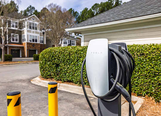 A Tesla charging station is located in front of a hedge and a building.