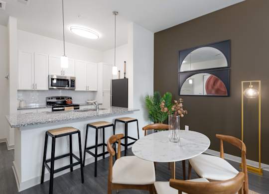 2-Bedroom Luxury Apartments in East Nashville, TN - Flats at Laurel and Pine - Dining Area with Vinyl Plank Flooring, Round Dining Table with Four Chairs, Floor Lamp, Breakfast Bar with Granite Countertop and Barstools, Pendant Lights, and View of Kitchen.