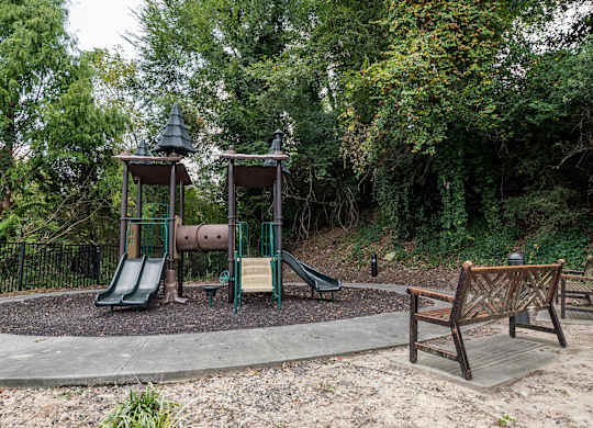 3-Bedroom Luxury Apartments in Atlanta, GA - The Eliza on Collier - Playground with a Multi-Play Structure on Wood Chips, Concrete Pathway, Wooden Bench, and Lush Green Trees.