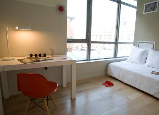 A red chair sits in front of a white desk in a room with a bed and a window.