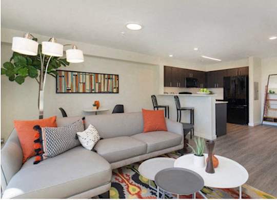 Living Room with Plank Flooring, Cozy Grey Couch, Nesting Tables, and Curved Lamp with Kitchen View at Arabella Apartment Homes, Washington
