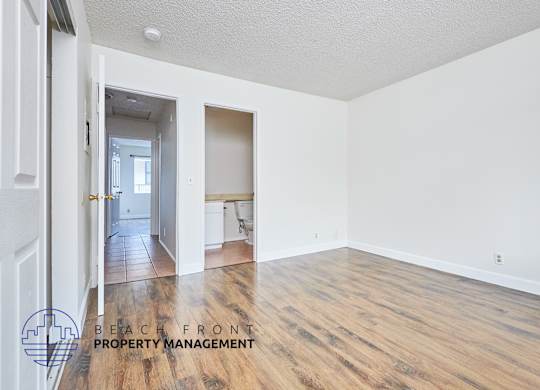 A room with wooden floors and white walls, with a logo for Beach Front Property Management in the bottom left corner.