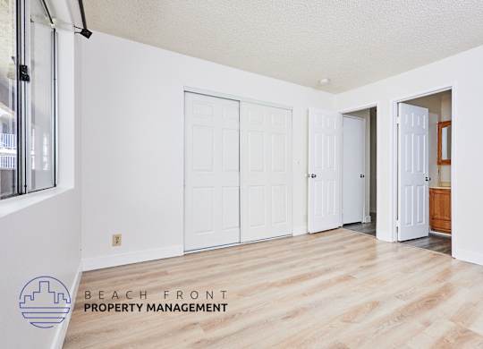 A room with white walls and wooden floors, with a logo for Beach Front Property Management in the bottom left corner.