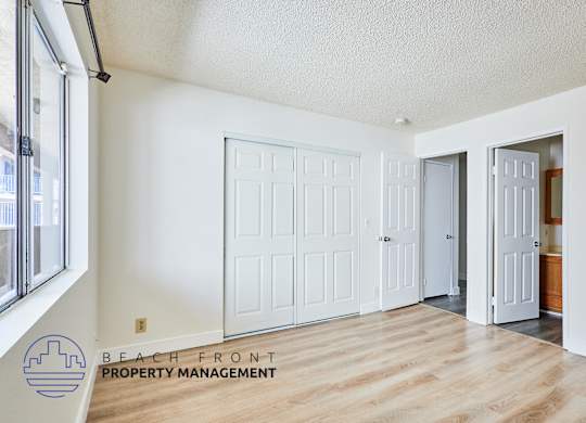 A room with white walls and wooden floors, featuring a white door and a window with a view of buildings outside.