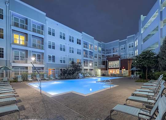 an outdoor swimming pool with chaise lounge chairs at night in front of an apartment building