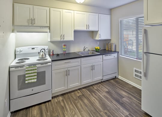 Kitchen with white cabinets and grey countertops. Image contains a white oven, sink and white dishwasher. One window and wood style flooring.at Woodhaven, Everett, Washington