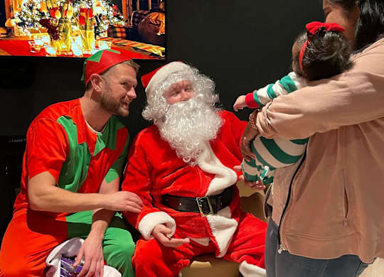 A woman is holding a child while two people dressed as elves sit with Santa Claus. at Cherrywood Apartments, D18 VY3V