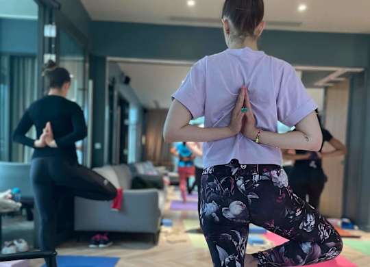 A woman in a purple shirt and floral pants is practicing yoga in a room with other people in the background. at Cherrywood Apartments, D18 VY3V