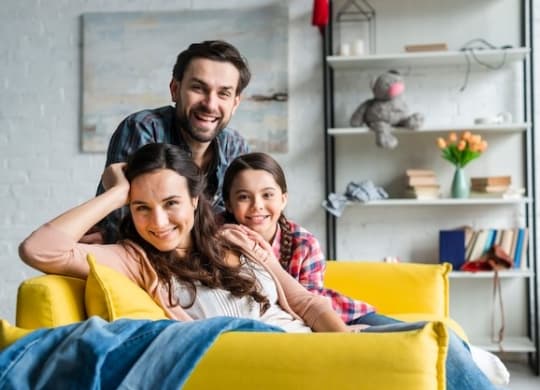 A family of three is sitting on a yellow couch in a room with a bookshelf in the background.