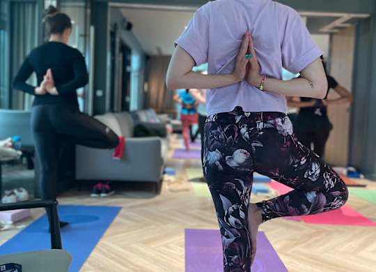 A woman in a purple shirt and floral pants is practicing yoga in a room with other people in the background at Cherrywood Apartments, D18 VY3V