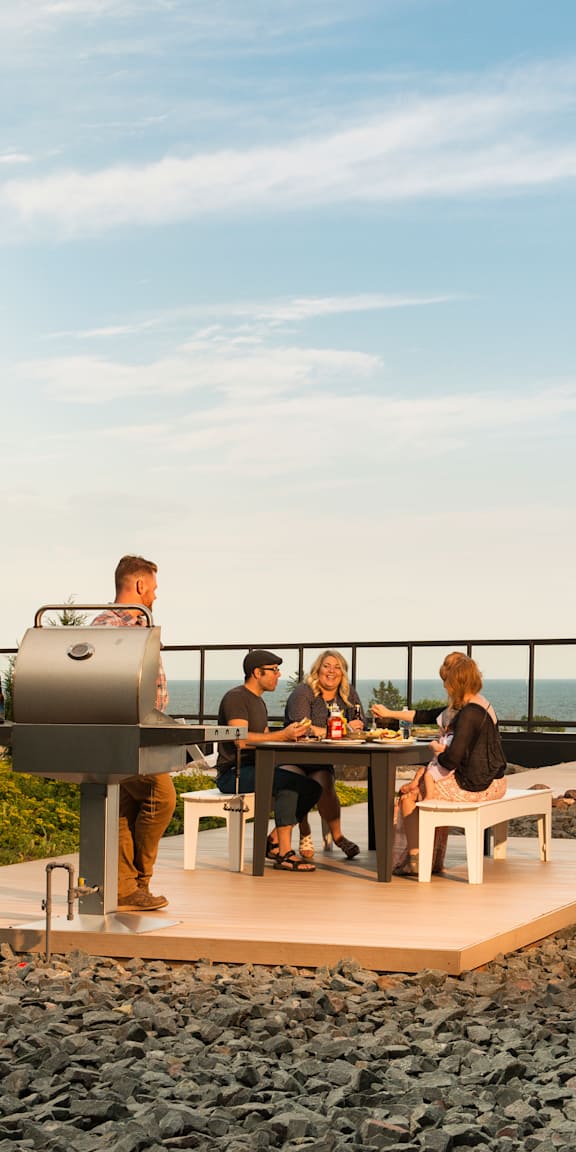 A group of people are gathered around a barbecue grill on a patio with a view of the ocean.