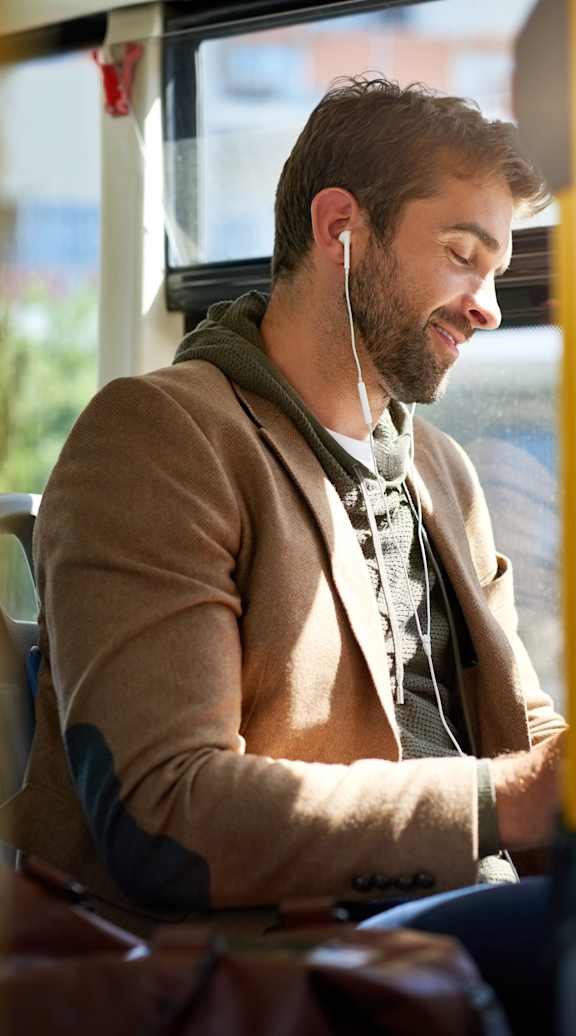 A man is sitting on a bus, wearing a brown jacket and holding a black tablet.