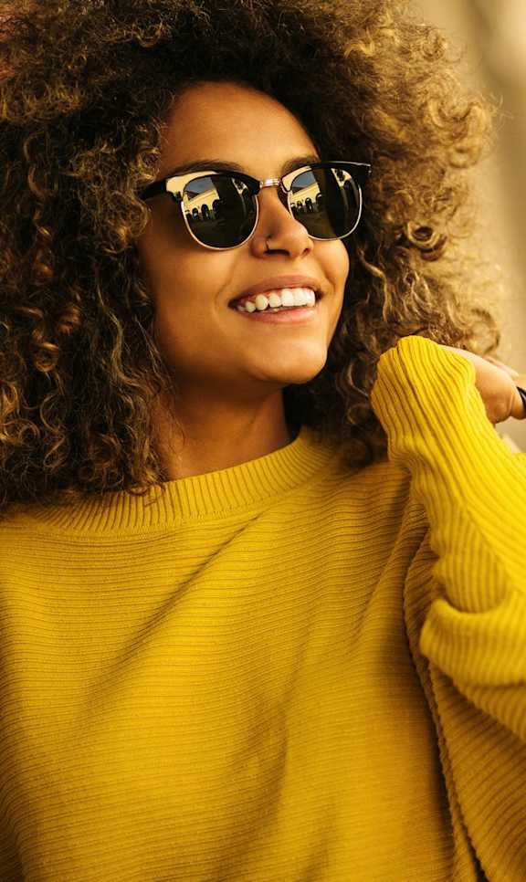 A woman with curly hair wearing sunglasses and a yellow sweater is holding shopping bags.