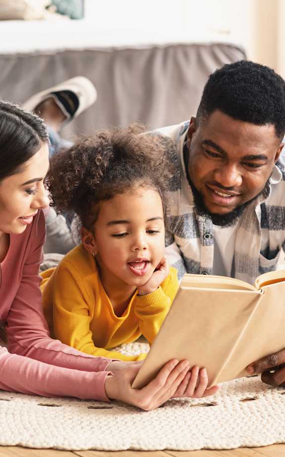 A family of three is reading a book together on the floor.