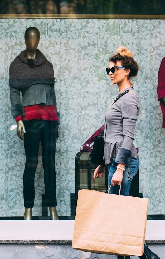 A woman with a shopping bag stands in front of a window display of mannequins.
