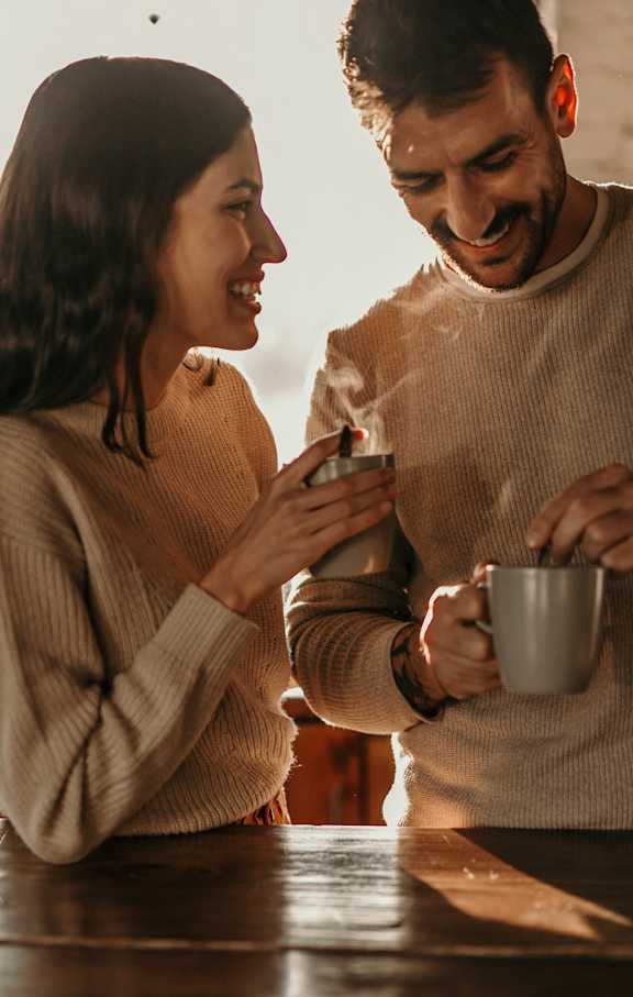 A man and a woman are standing at a table, with the man holding a mug and the woman holding a smartphone.