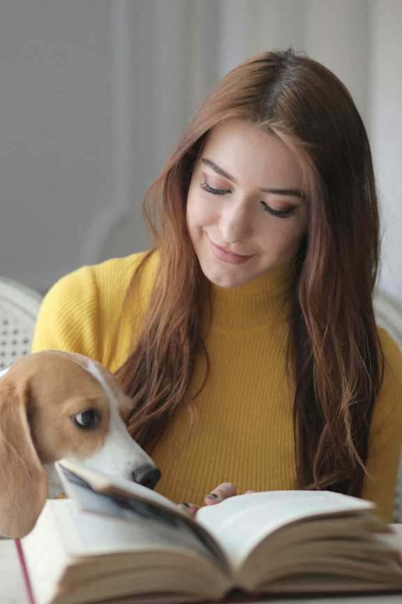 A woman in a yellow sweater is reading a book with a dog sitting beside her.