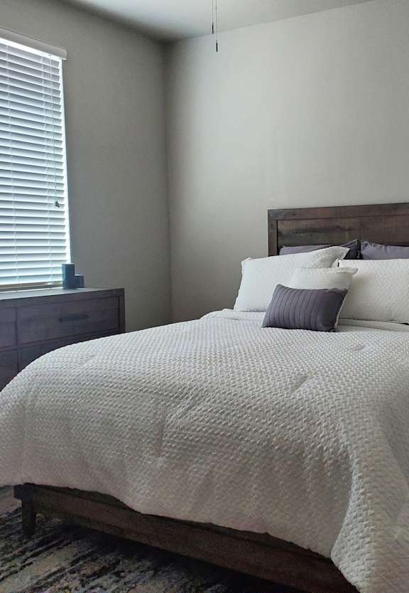 A bed with a white comforter and pillows sits in a room with a window and a door.