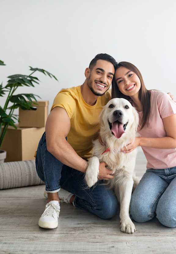 A man and woman are sitting on the floor with a dog between them, surrounded by cardboard boxes.