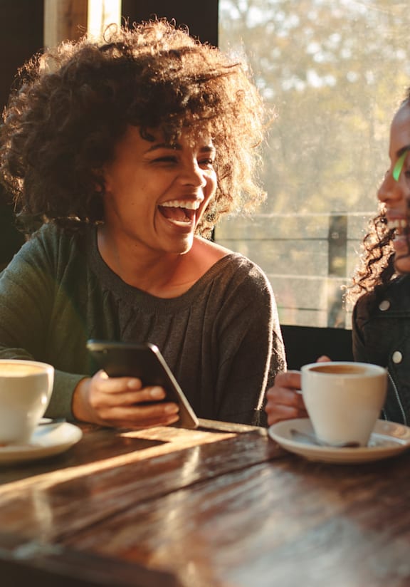 Two people are sitting at a table with cups of coffee and one is looking at their phone.
