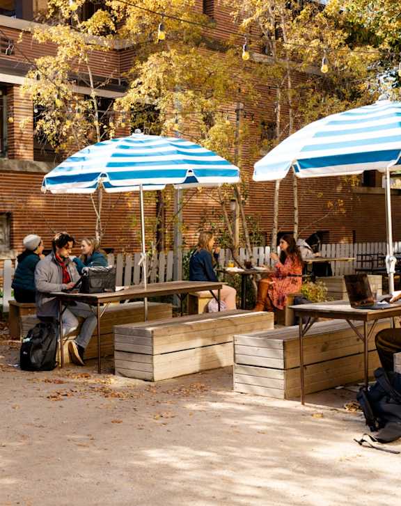 A group of people are sitting and standing on wooden benches under blue and white striped umbrellas.