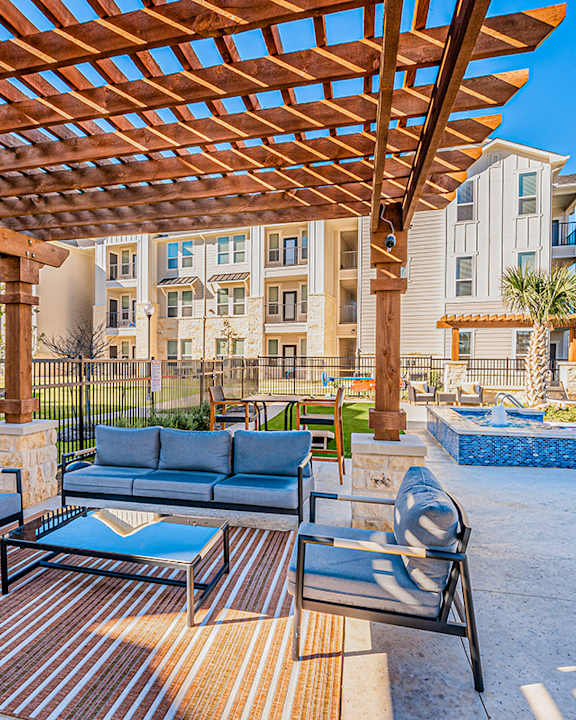 A wooden pergola shades a patio with a couch, chairs, and a pool.