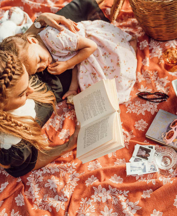 two women laying on a blanket reading a book at a picnic