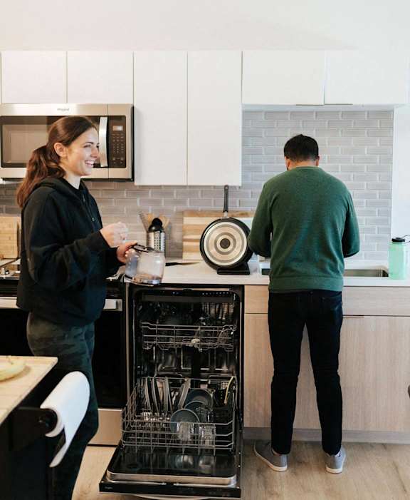 A man and a woman are standing in a kitchen with a dishwasher full of dishes.