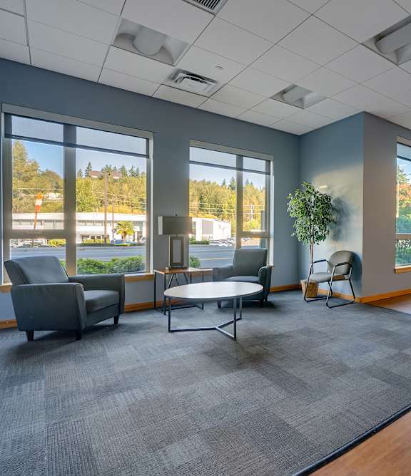 A spacious room with grey carpet and wooden flooring, furnished with chairs and tables.