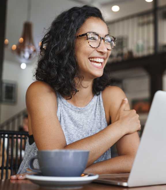 A woman with glasses is smiling while using a laptop at a table with a cup of coffee.