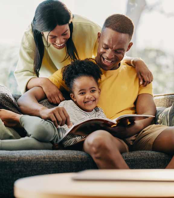 A family of three is sitting on a couch, with the child reading a book.